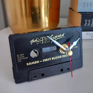 May include: A black cassette tape clock with white clock hands and a red second hand. The clock face features the text "The XMM Squad" and "RAMBO - FIRST BLOOD PART". A retro-style decorative item.