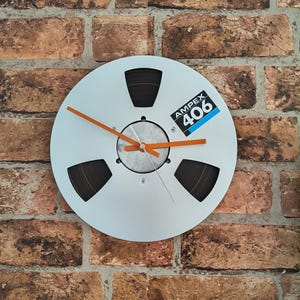 May include: A circular wall clock fashioned from a silver-coloured metal reel-to-reel tape spool. The clock features three orange hands and a central label displaying the text AMPEX 406 in white and blue against a black background.
