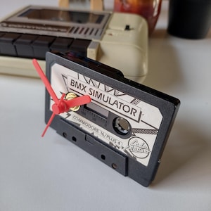 May include: A black cassette tape clock with red hands. The clock face displays "BMX SIMULATOR" and "COMMODORE 16/PLUS 4". This clock is a distinctive decorative piece, ideal for a retro or gaming-themed room.
