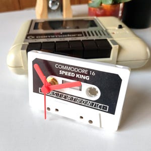 May include: A white cassette tape clock with red hands, featuring the text "COMMODORE 16 SPEED KING" and "MASTERTRONIC". A vintage beige Commodore 16 computer is in the background.