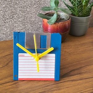 May include: A blue floppy disk repurposed as a clock with bright yellow hour, minute, and second hands. The disk features a white label area with red horizontal lines and the word INDEX printed in red text near the top left corner of the label.