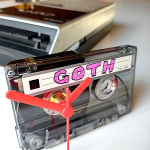 May include: A clear cassette tape clock with red hands. The label reads "GOTH" in pink letters. A vintage cassette player is in the background. The clock is a unique decorative item.