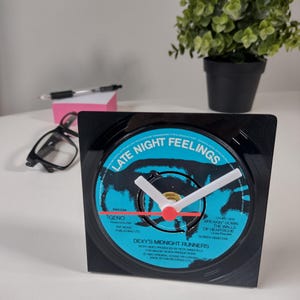 May include: A black square clock with a vinyl record face. The clock face is blue and black with white clock hands and red accents. The text "LATE NIGHT FEELINGS" is visible on the clock face.