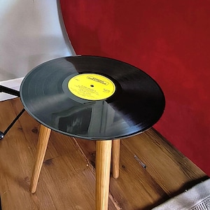 May include: A small side table featuring a circular top made from a black vinyl record with a yellow centre label that reads Stereo Gold Award. The table is supported by three light-coloured wooden legs that taper towards the floor.