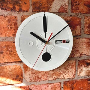 May include: A white wall clock with black hands and a red second hand, mounted on a brick wall. The clock face has a circular design with the text "Scotch 3M". The clock's design resembles a reel of tape.
