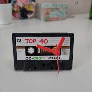 May include: A black cassette tape clock with red clock hands. The tape reads "TOP 40" in red lettering. The clock is a TDK OD-C90 model.