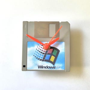 May include: A square grey floppy disk repurposed as a wall clock. The centre features a label with the Microsoft Windows 95 logo, including the blue sky background and colourful window icon. Bright orange clock hands are attached to the centre.