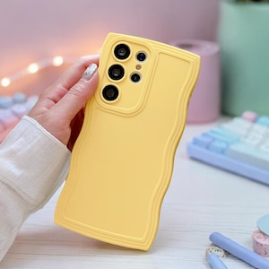 Puede incluir: Funda de teléfono amarilla con un diseño de borde ondulado. La funda tiene tres recortes para la cámara y un recorte para el flash. La funda se sostiene en una mano, con un teclado y otros accesorios de escritorio en el fondo.