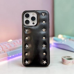 Puede incluir: Funda de teléfono negra con estrellas y tachuelas plateadas. La funda tiene una apariencia texturizada y un recorte para la cámara. Las estrellas están espaciadas uniformemente, agregando un toque decorativo al accesorio.