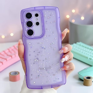 Puede incluir: Una funda de tel&eacute;fono lavanda transparente con un dise&ntilde;o de borde ondulado. La funda est&aacute; llena de purpurina y detalles en forma de estrella. La funda del tel&eacute;fono tiene un logotipo de Samsung en la parte inferior.