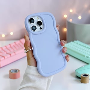 Puede incluir: Funda de teléfono lila con un borde ondulado. La funda tiene una superficie lisa y se muestran las lentes de la cámara del teléfono. La funda se sostiene en una mano, con un teclado rosa y otros suministros de oficina en el fondo.