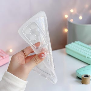 Puede incluir: Funda de tel&eacute;fono transparente y flexible con detalles de purpurina. La funda se sostiene en una mano, mostrando su dise&ntilde;o transparente y el recorte de la c&aacute;mara. La funda parece estar hecha de un material suave y flexible.