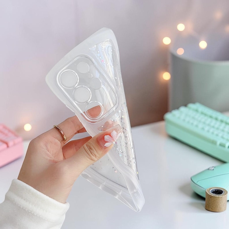 Puede incluir: Funda de tel&eacute;fono transparente y flexible con detalles de purpurina. La funda se sostiene en una mano, mostrando su dise&ntilde;o transparente y el recorte de la c&aacute;mara. La funda parece estar hecha de un material suave y flexible.
