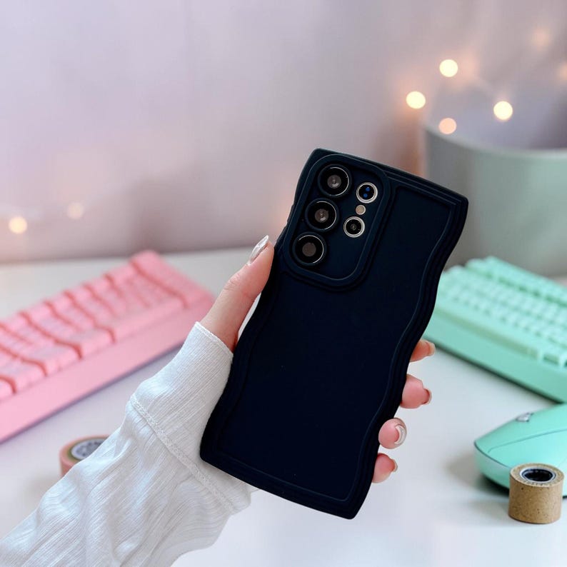 Peut inclure: Une coque de t&eacute;l&eacute;phone bleu marine avec un design &agrave; bord ondul&eacute;. L'&eacute;tui a une d&eacute;coupe pour l'appareil photo avec trois lentilles et deux cercles plus petits. L'&eacute;tui est tenu dans une main, avec un clavier rose et un clavier vert menthe en arri&egrave;re-plan.
