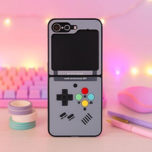 Puede incluir: Funda de teléfono gris diseñada para parecerse a una consola de juegos portátil retro. La funda presenta una pantalla, un mando direccional y botones de colores. El texto "START GAME" y "GAME DOYA3VENGE SP" es visible.