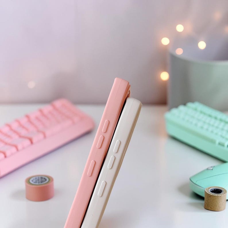 Peut inclure: Deux coques de t&eacute;l&eacute;phone, une rose et une blanc cass&eacute;, sont appuy&eacute;es l'une contre l'autre. Les coques ont des d&eacute;tails de boutons sur le c&ocirc;t&eacute;. Un clavier rose, un rouleau de ruban adh&eacute;sif et un clavier et une souris vert menthe sont en arri&egrave;re-plan.
