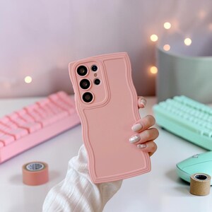 Peut inclure: Une coque de t&eacute;l&eacute;phone rose clair avec un bord ondul&eacute; et textur&eacute;. L'&eacute;tui comporte des d&eacute;coupes pour les objectifs de l'appareil photo et les boutons du t&eacute;l&eacute;phone. L'arri&egrave;re-plan comprend un clavier rose, un clavier vert menthe et un rouleau de ruban adh&eacute;sif.