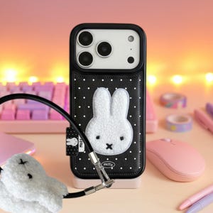 May include: Black phone case with a white bunny design. The case has a black strap and a white fluffy bunny charm. The case is on a pink stand, with a pink mouse and pens in the background.