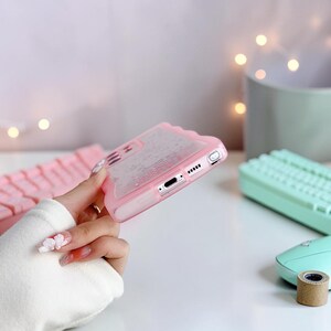 Puede incluir: Una funda de tel&eacute;fono rosa con un borde ondulado se sostiene. La funda es transparente con purpurina en el interior. La funda del tel&eacute;fono tiene recortes para la c&aacute;mara, el puerto de carga y los botones. Otros elementos en el fondo incluyen un teclado y un rat&oacute;n.