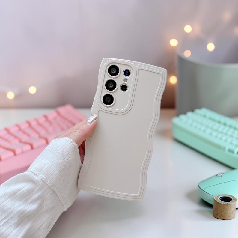 Peut inclure: Coque de t&eacute;l&eacute;phone blanche avec un bord ondul&eacute;. Le bo&icirc;tier comporte une d&eacute;coupe pour l'appareil photo avec trois objectifs et un flash. Le bo&icirc;tier est tenu par une personne. Un clavier rose, un clavier vert menthe et une souris vert menthe sont en arri&egrave;re-plan.