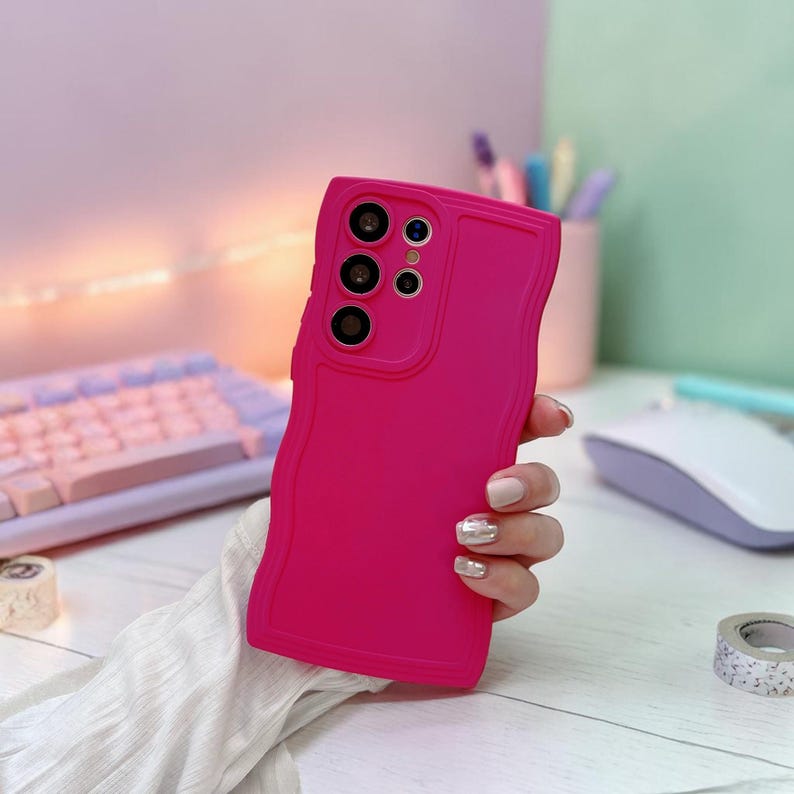 Peut inclure: Une coque de t&eacute;l&eacute;phone rose vif avec un bord ondul&eacute;. L'&eacute;tui pr&eacute;sente des d&eacute;coupes pour les objectifs de l'appareil photo et les boutons du t&eacute;l&eacute;phone. La coque est tenue dans une main avec des ongles manucur&eacute;s.