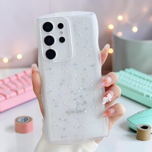 Puede incluir: Funda transparente para tel&eacute;fono con borde ondulado, llena de purpurina y detalles en forma de estrella. La funda tiene un recorte para la c&aacute;mara y el logotipo de Samsung. La funda la sostiene una persona con u&ntilde;as largas.