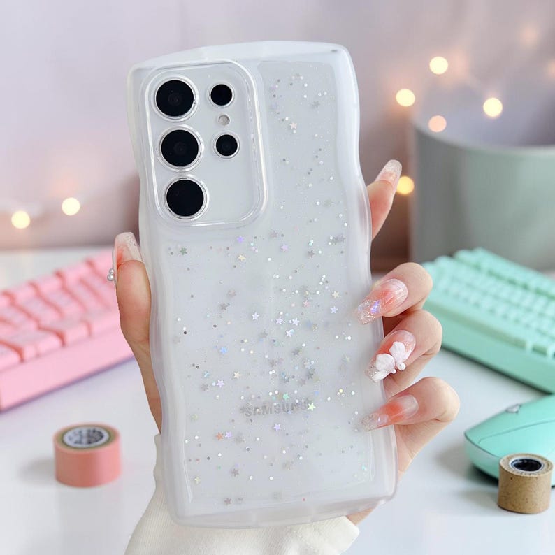 Puede incluir: Funda transparente para tel&eacute;fono con borde ondulado, llena de purpurina y detalles en forma de estrella. La funda tiene un recorte para la c&aacute;mara y el logotipo de Samsung. La funda la sostiene una persona con u&ntilde;as largas.