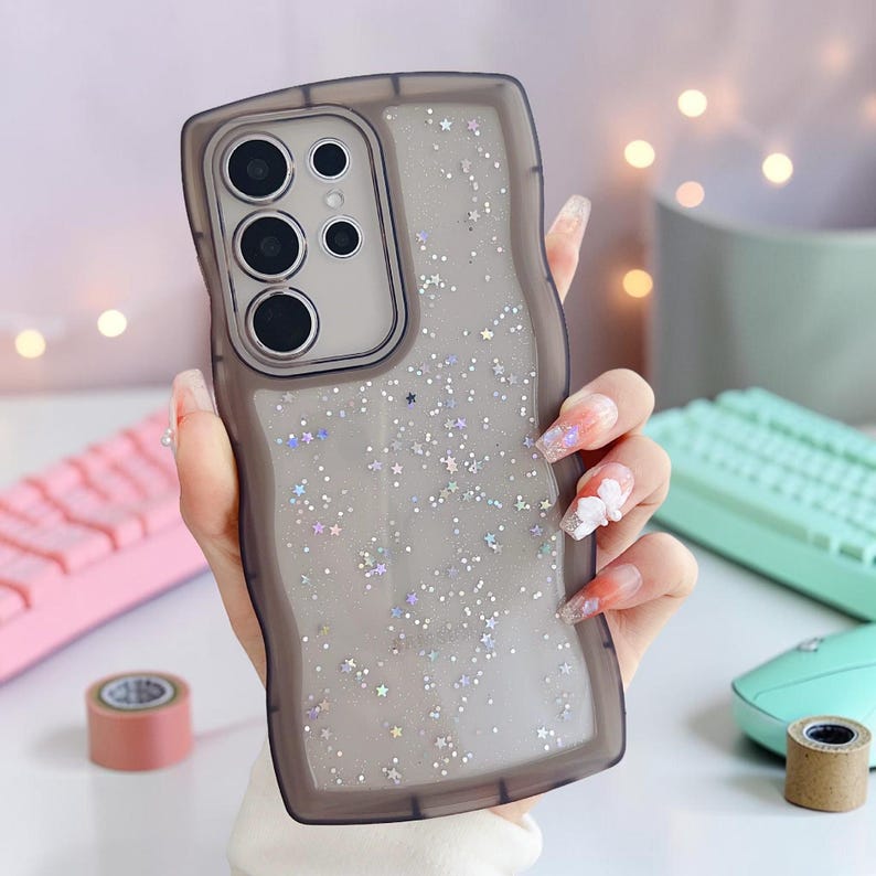 Puede incluir: Una funda transparente para tel&eacute;fono con un borde ondulado de color gris oscuro. La funda est&aacute; llena de purpurina plateada y detalles en forma de estrella. La funda del tel&eacute;fono tiene un m&oacute;dulo de c&aacute;mara con tres lentes.