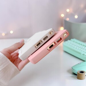 Peut inclure: Deux coques de t&eacute;l&eacute;phone, une blanche et une rose, sont empil&eacute;es. La coque blanche a un motif textur&eacute;, tandis que la coque rose est lisse. Les deux coques ont des ouvertures pour le port de charge et l'appareil photo.