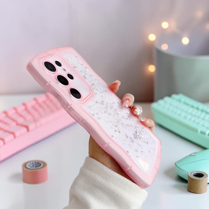 Puede incluir: Una funda de tel&eacute;fono rosa con un centro transparente lleno de purpurina plateada. La funda tiene un borde elevado y un recorte para la c&aacute;mara. La funda se sostiene frente a un fondo borroso.