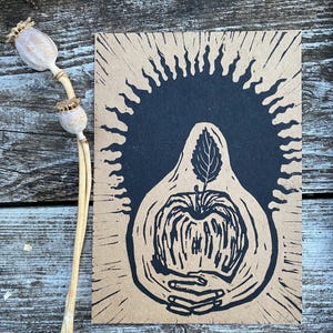 May include: A linocut print on tan paper features a black apple with a leaf, held by hands, surrounded by a sunburst design. The print is next to dried poppy seed pods on a wooden surface. The artwork is a unique piece.