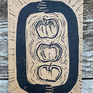 Könnte beinhalten: Ein rechteckiger, handgefertigter Druck mit drei stilisierten Äpfeln in einem schwarzen Ovalrahmen. Der Druck befindet sich auf einem beigefarbenen Hintergrund mit radialen Linien um den Rahmen, was eine rustikale, künstlerische Ästhetik erzeugt.