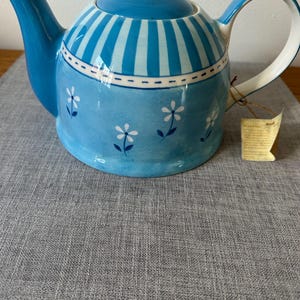 May include: A blue ceramic teapot with a white handle and lid. The teapot has a striped pattern on the lid and a floral design on the body. The teapot is on a grey surface.