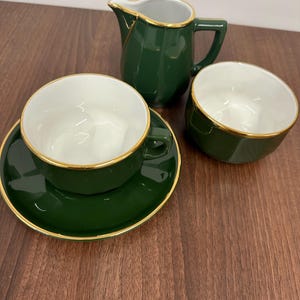 May include: A green tea set with gold edging. The set comprises a teacup and saucer, a creamer jug, and a sugar bowl. The inside of the teacup, creamer, and sugar bowl are white. The tea set is on a wooden surface.