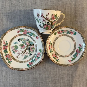 May include: A white teacup and two saucers with a floral design. The teacup and saucers have a gold trim. The floral design includes pink and green flowers and branches. The teacup has a gold handle.