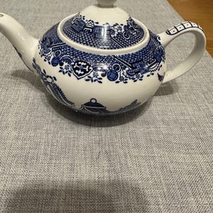 May include: A white ceramic teapot with a blue and white floral design. The teapot features a rounded body, a curved spout, and a handle with a geometric pattern. The lid is topped with a small, round knob.