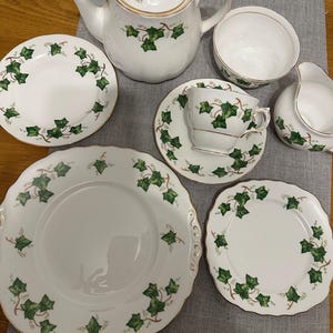 May include: A white porcelain tea set featuring a green ivy leaf pattern and gold edging. The set comprises a teapot, teacups, saucers, plates, a sugar bowl, and a creamer. The plates have decorative, scalloped edges.