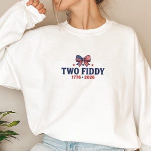 May include: A white crewneck sweatshirt featuring an embroidered design on the chest. The graphic includes a bow with an American flag pattern, the text "TWO FIDDY" in blue, and the years "1776 • 2026" in red, accented by two small red stars.