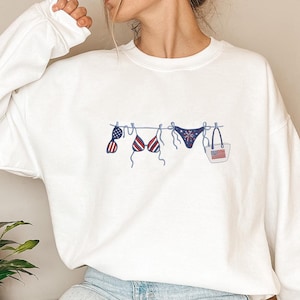 May include: White sweatshirt featuring an embroidered design of a bikini and beach bag. The bikini top, bottom, and bag are decorated with red, white, and blue patterns, including the American flag. The design is a patriotic theme.