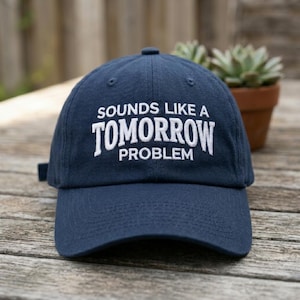 May include: A navy blue baseball cap featuring white embroidered text on the front that reads "SOUNDS LIKE A TOMORROW PROBLEM". The cap has a structured crown, a curved brim, and a fabric strap with a metal buckle closure at the back.