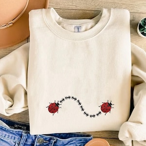 Può includere: Felpa color crema con un design stravagante di due coccinelle rosse collegate da una fila di piccole formiche nere. Le coccinelle hanno macchie nere e antenne. La felpa è piegata ordinatamente su una superficie di legno.