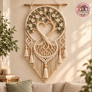 Könnte beinhalten: Ein Makramee-Wandbehang mit einem runden Holzrahmen und einem Baummotiv. Der Baum besteht aus geflochtenen Seilzweigen, die eine Herzform bilden, und ist mit kleinen weißen Blüten sowie grünen Blättern verziert. Das Stück enthält Quasten und Holzperlen.