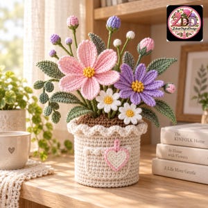 Puede incluir: Un arreglo floral de ganchillo con flores rosas, moradas y blancas con hojas verdes en una maceta color crema. La maceta tiene un borde superior festoneado y un pequeño parche rosa en forma de corazón en la parte delantera.
