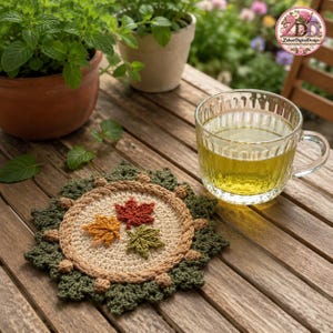 May include: A round crochet coaster featuring a beige centre decorated with three small maple leaves in red, orange, and green. The coaster has a dark green leafy border with small tan accents. A clear glass mug filled with light green tea sits nearby.