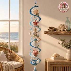May include: A vertical hanging spiral mobile featuring crocheted ocean elements including a seashell, a red starfish, a seahorse, a jellyfish, red coral, and a whale tail, accented with blue and white beads and a circular logo at the top right.