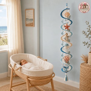 May include: A vertical hanging mobile featuring a spiral design in shades of blue and cream, decorated with crocheted sea life elements including a seashell, starfish, seahorse, jellyfish, coral, and a whale tail, finished with a tassel at the bottom.