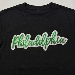 May include: Black crew neck t-shirt with the word "Philadelphia" in a green, glittery script font outlined in white. The shirt is a solid color, and the text is centered.