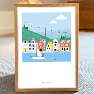 Könnte beinhalten: Ein gerahmter Kunstdruck mit einer farbenfrohen Illustration einer europäischen Stadt am Fluss. Das Kunstwerk zeigt ein Segelboot, Seilbahnen und den Namen der Stadt, Rudesheim, am unteren Rand. Der Rahmen ist aus hellem Holz.