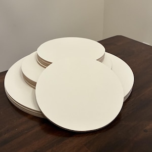 May include: A stack of multiple circular, flat, off-white wooden boards of varying diameters. The boards have smooth, light-colored surfaces and visible layered edges, arranged in a tiered pile with the smallest discs placed on top.