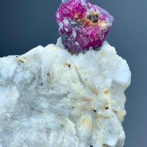 Pode incluir: Uma imagem de perto de um cristal de rubi bruto em cima de uma formação mineral branca. O rubi é rosa vibrante com toques de azul e dourado. O mineral branco tem uma superfície áspera e texturizada.